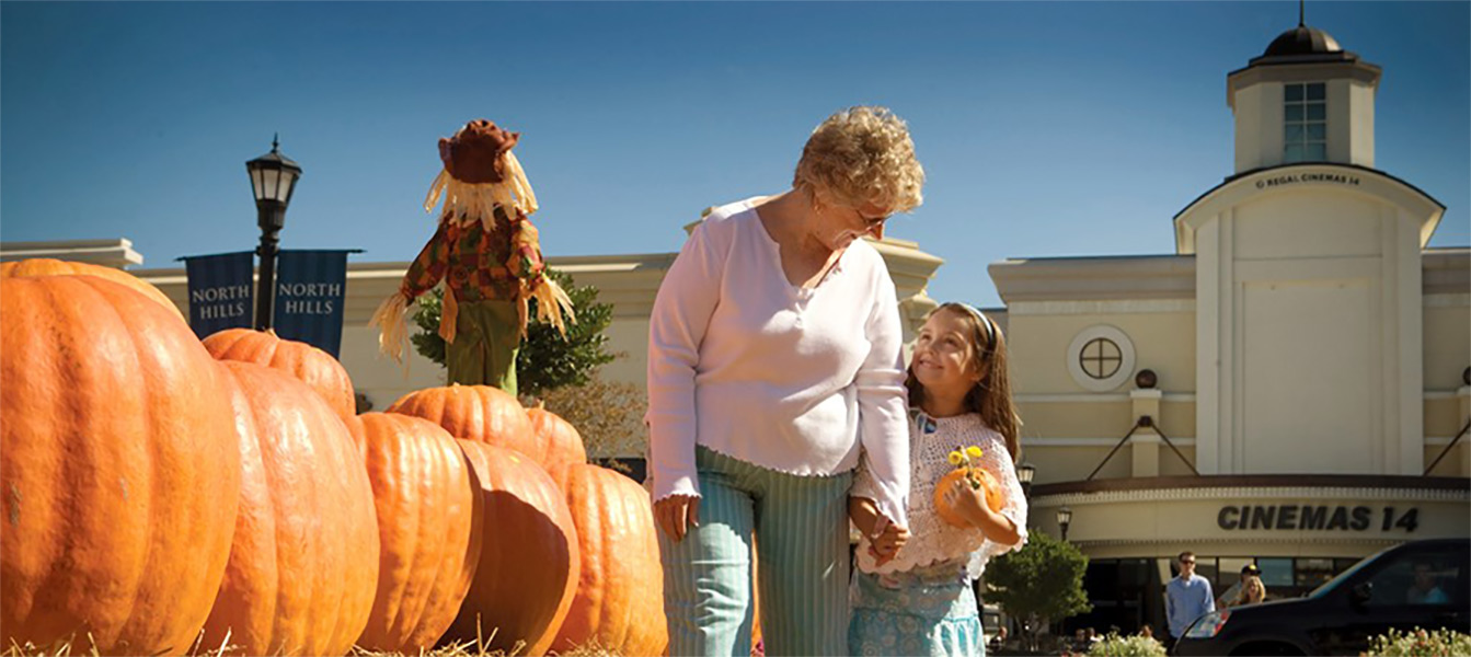 Older woman and young girl smile at each other and hold hands while walking next to a pumpkin patch in the North Hill lawn.