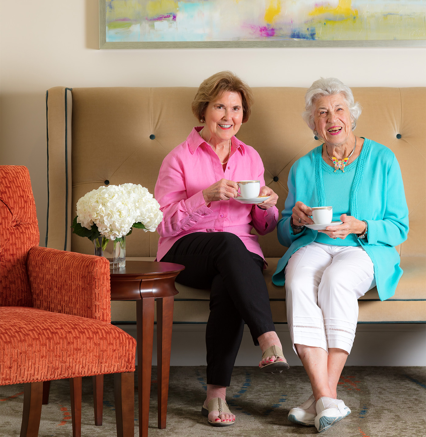Two female residents at The Cardinal sitting on a couch enjoying some coffee and smiling.
