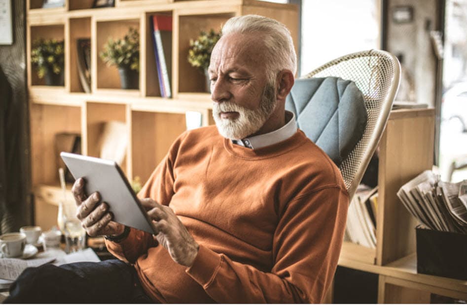 Older gentleman with a beard sitting in a chair looking at something on a tablet computer.