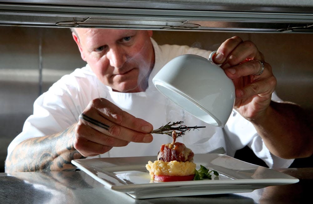 Chef delicately putting a sprig of rosemary on a plate of some very fancy looking food and preparing to put a cloche over it.