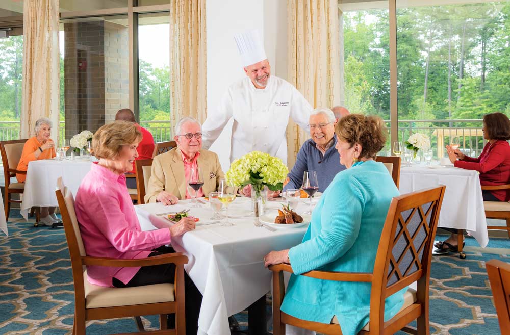 Residents smiling and enjoying dinner while they talk with the chef at the table.
