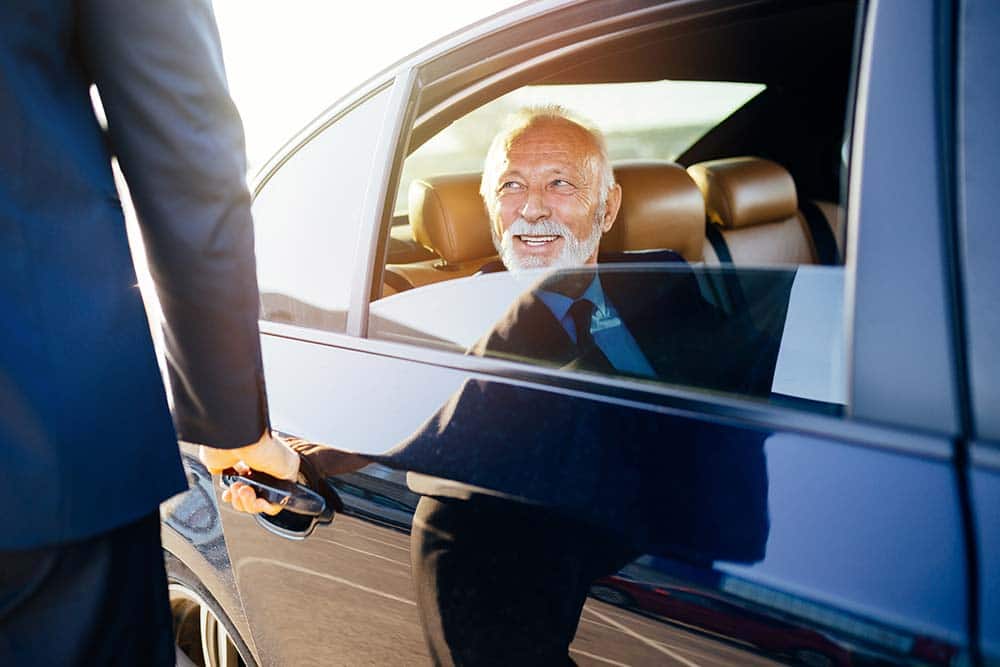 Smiling gentleman sitting in the backseat of a black sedan smiling while an unseen valet reaches to open the door.