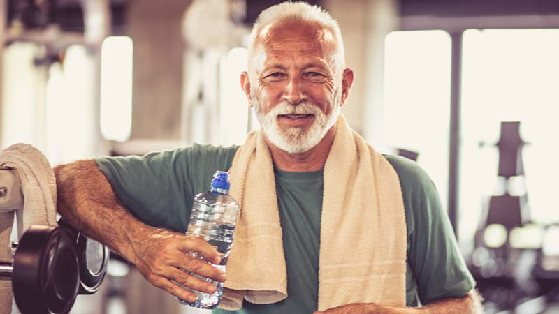 Man with a beard and towel draped around his neck holding a water bottle and smiling in a gym.
