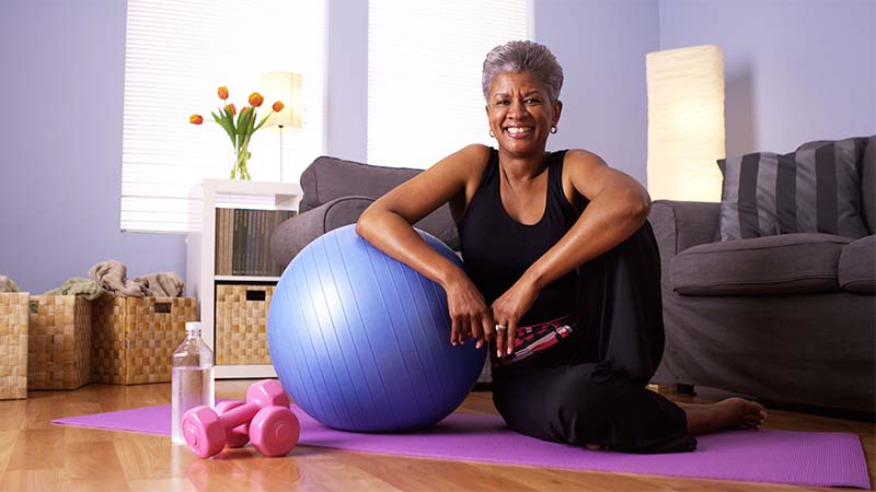 Smiling woman in workout attire smiling and sitting on the floor with a yoga mat, weights, and an exercise ball.