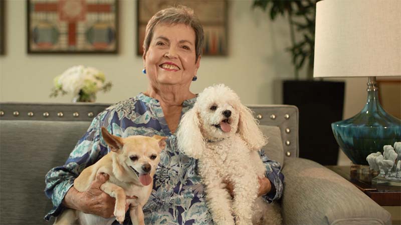 Deanna, Cardinal Resident, sitting on her couch and smiling while holding her two dogs.