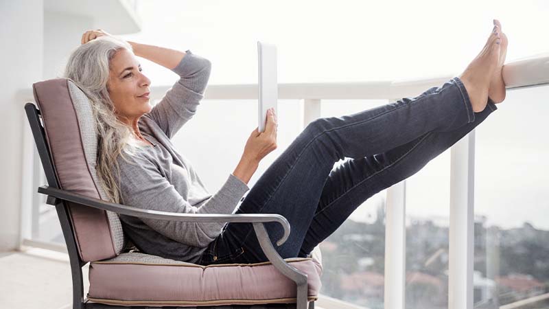 Woman in jeans and a sweater reclining with her feet up on the railing of her balcony while looking at a tablet computer.