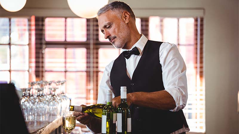 Waiter in vest and bow tie pouring glasses of wine at the bar.